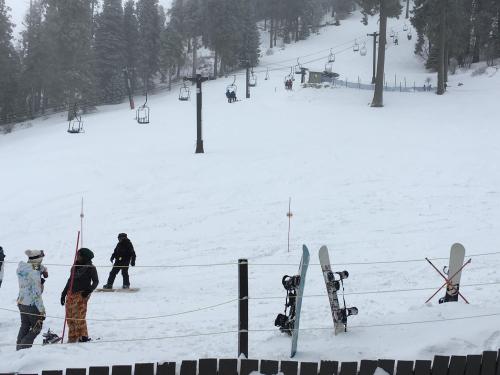A snowy ski resort scene with snowboarders preparing and ski lifts in the background among tall trees.