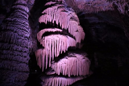 A cave formation with large, pinkish stalactites and stalagmites illuminated by soft purple light.