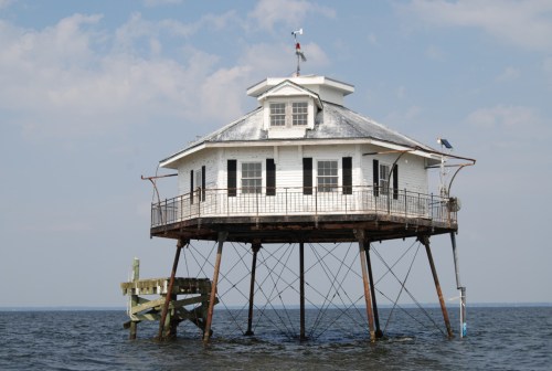 A stilted white house on water, featuring a balcony and a weather vane, under a blue sky with scattered clouds.