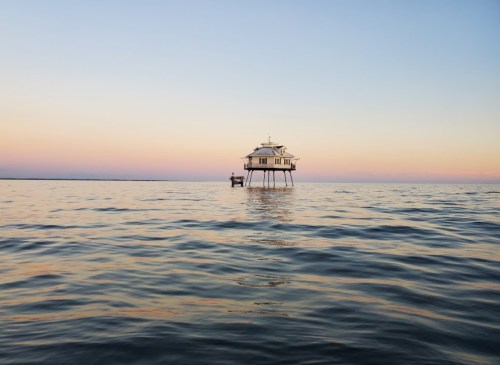 A serene waterscape featuring a stilted house on calm water during sunset, with soft pastel colors in the sky.