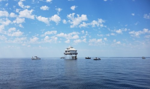 A scenic view of a stilted building on calm water, surrounded by boats under a blue sky with scattered clouds.