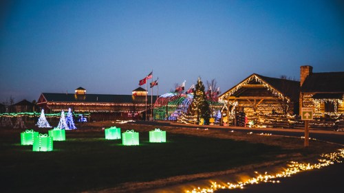 Colorful holiday lights illuminate a festive outdoor scene with decorated trees and gift-shaped displays at dusk.