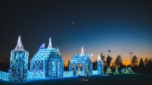A winter scene featuring illuminated structures and trees in blue and green lights against a twilight sky.