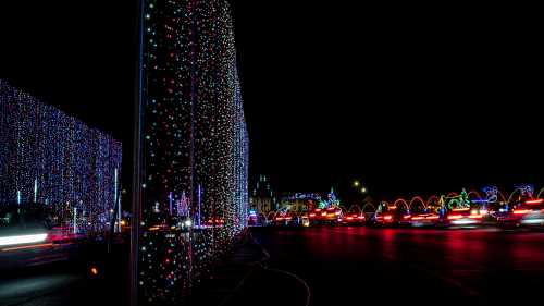 Colorful holiday lights illuminate a street at night, with cars passing by and festive decorations in the background.