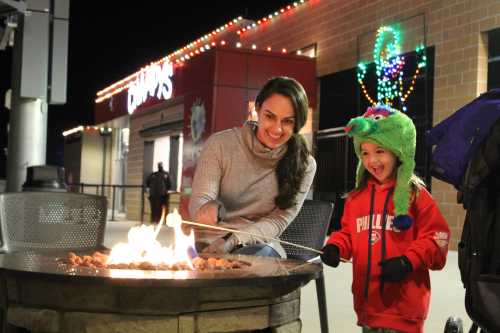 A woman and a child in a green hat roast marshmallows by a fire pit, smiling in a festive outdoor setting.