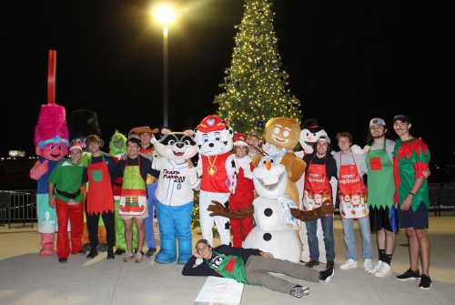 A group of people in festive costumes poses in front of a Christmas tree at night, celebrating the holiday season.