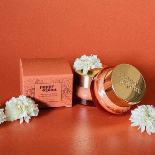 A jar of Poppy & Pout lip scrub next to its box, surrounded by white flowers on an orange background.