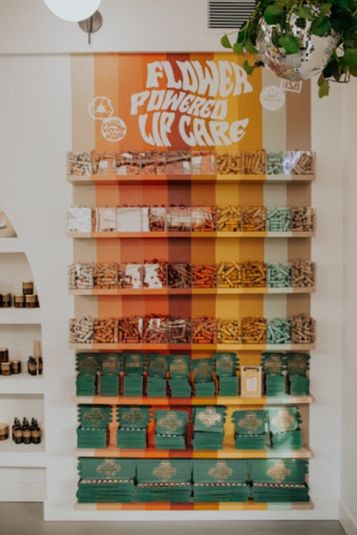 A colorful display of various beauty products arranged in a gradient on wooden shelves, featuring vibrant packaging.