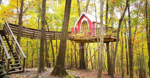 A charming red treehouse with a deck, nestled among vibrant autumn trees in a forest.