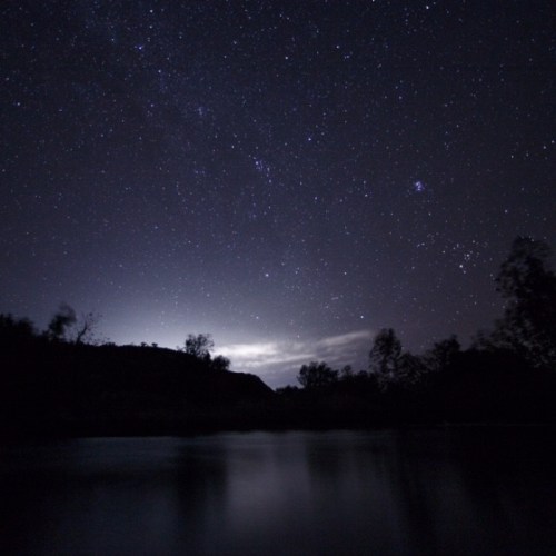 A serene night sky filled with stars above a calm body of water, with silhouettes of trees and hills in the foreground.