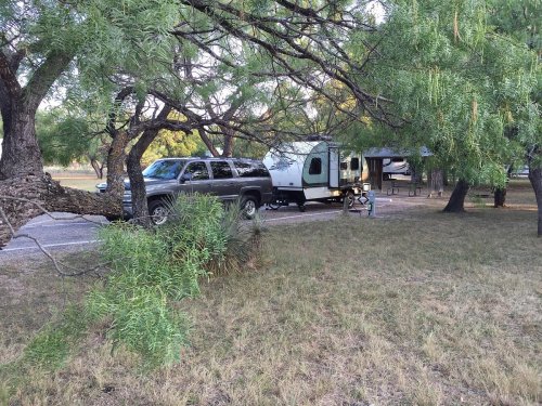 A camper and SUV parked under trees in a grassy area, with another camping setup visible in the background.