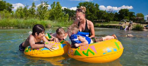 A woman and three children play in a river on colorful inner tubes, enjoying a sunny day outdoors.