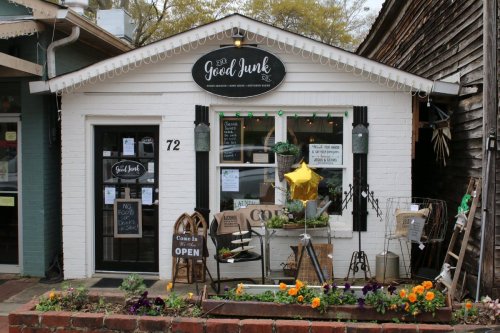 A quaint shopfront with a sign reading "Good Junk," featuring plants, flowers, and various decorative items outside.