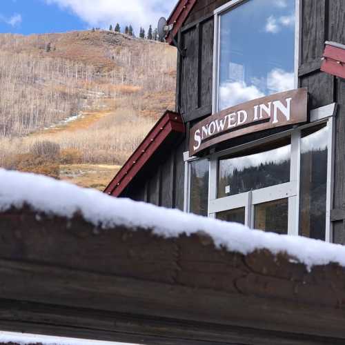 A wooden building with a sign reading "Snowed Inn," surrounded by snow and mountains under a clear blue sky.