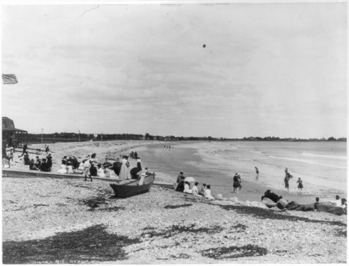 A black and white photo of a beach scene with people relaxing, walking, and playing near the water.