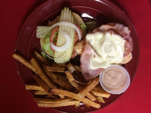 A plate with a sandwich topped with ham, lettuce, tomato, pickles, and mayonnaise, served with fries and dipping sauce.