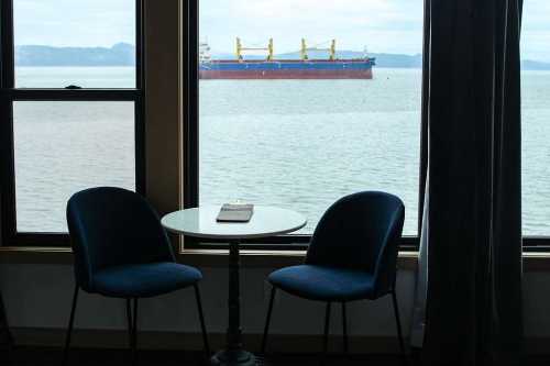 A view from a window featuring two chairs and a table, with a large cargo ship in the water outside.