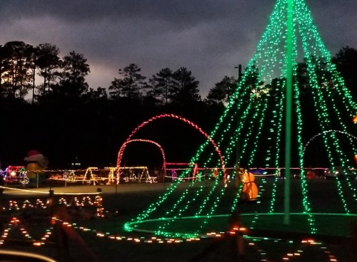 Colorful holiday lights illuminate a park, featuring a large tree and festive decorations against a darkening sky.