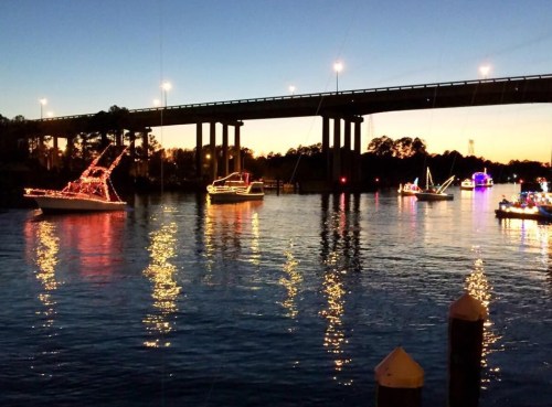 Boats decorated with lights float on a river at dusk, with a bridge illuminated in the background.