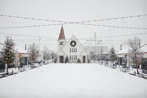 A snowy scene of a quaint building with a spire, surrounded by trees and benches, decorated for the winter season.
