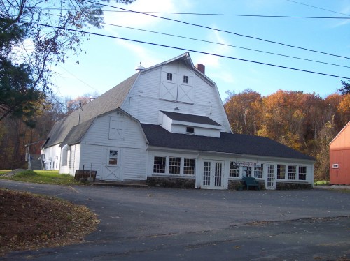 A large white barn with a dark roof, surrounded by trees with autumn foliage and a gravel driveway.