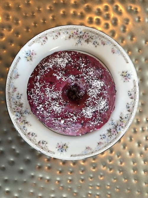 A purple frosted donut with powdered sugar on a decorative plate, set against a textured metallic background.