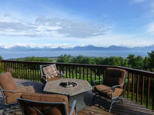 A scenic view from a deck with chairs and a fire pit, overlooking mountains and a body of water under a blue sky.
