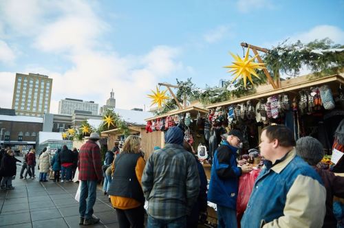 Crowd at a festive market with wooden stalls decorated with stars, selling handmade goods against a city skyline.