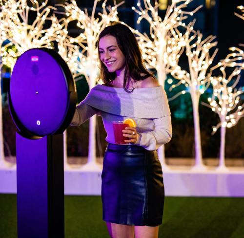 A woman in a stylish outfit smiles while interacting with a drink dispenser, surrounded by illuminated trees.