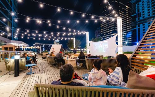 A rooftop lounge at night with people sitting around, string lights overhead, and a movie screen in the background.