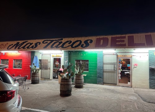A colorful taco deli at night, featuring green and red walls, cacti in barrels, and outdoor seating.
