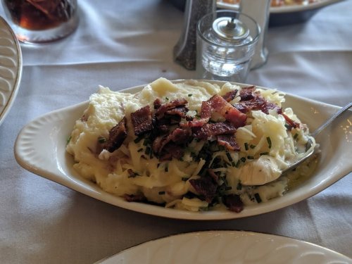 A dish of creamy mashed potatoes topped with crispy bacon and chives, served in a white oval bowl.