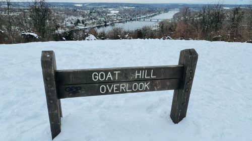 A wooden sign reading "Goat Hill Overlook" in a snowy landscape with a river and hills in the background.