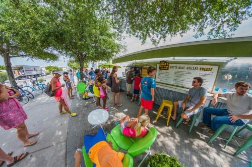 A busy food truck scene with people waiting in line, enjoying outdoor seating under trees on a sunny day.