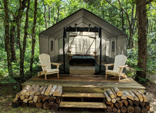 A cozy tent on a wooden deck in a forest, with two chairs and stacked logs in front.
