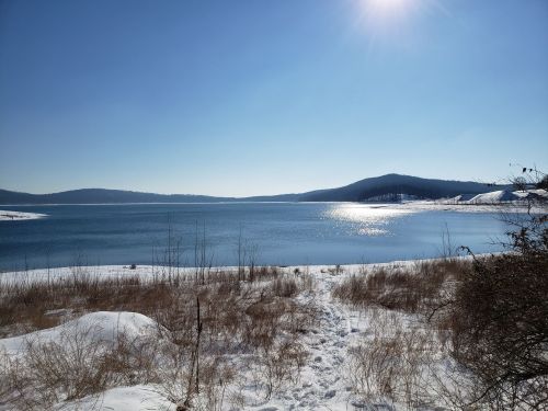 A serene winter landscape featuring a snowy shore, calm lake, and distant hills under a clear blue sky.