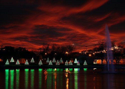 Colorful Christmas trees illuminated by lights reflect in a calm water body under a dramatic sunset sky.