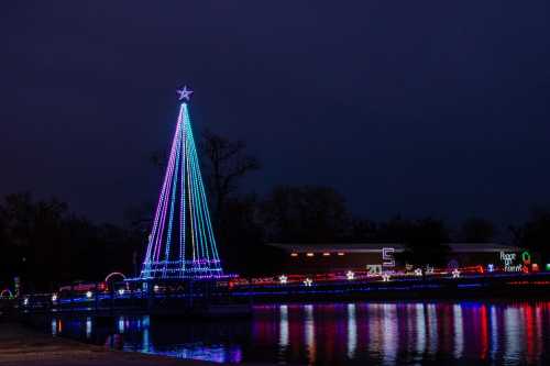 A brightly lit Christmas tree with colorful lights stands by a lake, reflecting in the water at dusk.