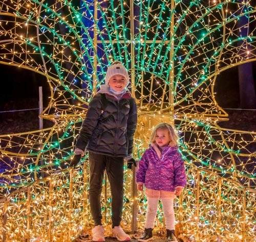 Two children stand in front of a large, illuminated holiday display made of colorful lights.