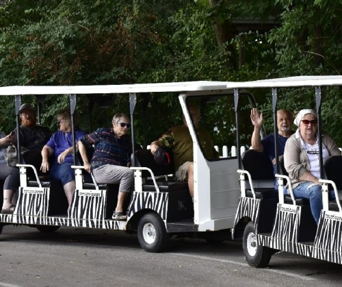 A group of people sitting in a tram-like vehicle, surrounded by greenery, some waving and smiling at the camera.