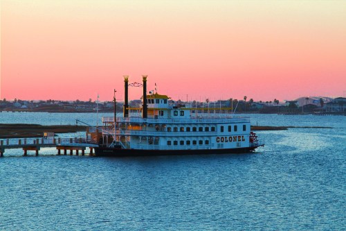 A riverboat named "Colonel" docked on calm waters during sunset, with a colorful sky in the background.