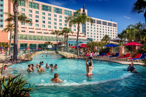 A vibrant hotel pool scene with people swimming, lounging, and palm trees under a clear blue sky.