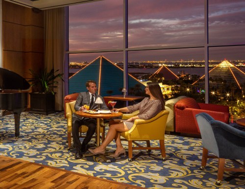 A couple enjoys drinks at a stylish lounge with a view of pyramids and a sunset in the background.