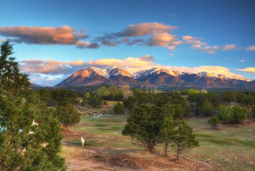 Scenic landscape featuring snow-capped mountains, green trees, and a colorful sky at sunset.
