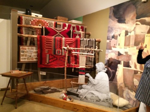 A museum display featuring traditional textiles, a weaving figure, and a painted backdrop of a desert landscape.