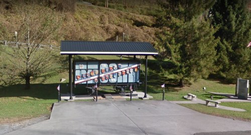 A bus stop shelter with a schedule display, surrounded by trees and a paved area, in a rural setting.