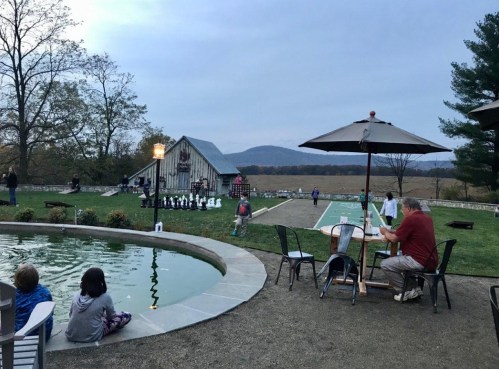 A scenic outdoor area with a pond, chess set, and people enjoying the evening near a barn and mountains in the background.
