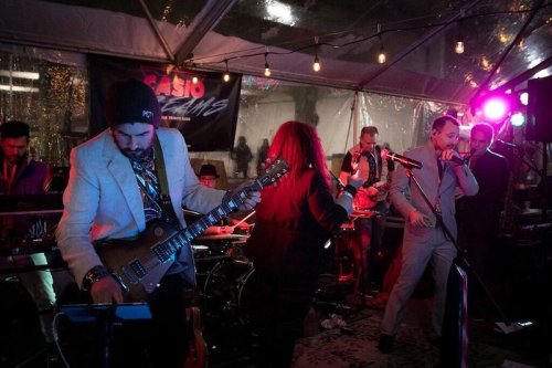A lively band performs under a tent with colorful lights, featuring musicians playing instruments and singing.