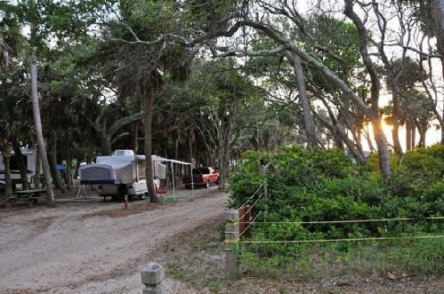 A serene campsite with trailers and boats surrounded by lush trees at sunset.