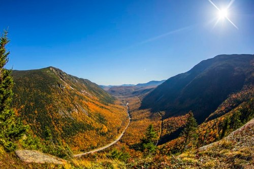 A panoramic view of a valley surrounded by mountains, showcasing vibrant autumn foliage under a clear blue sky.
